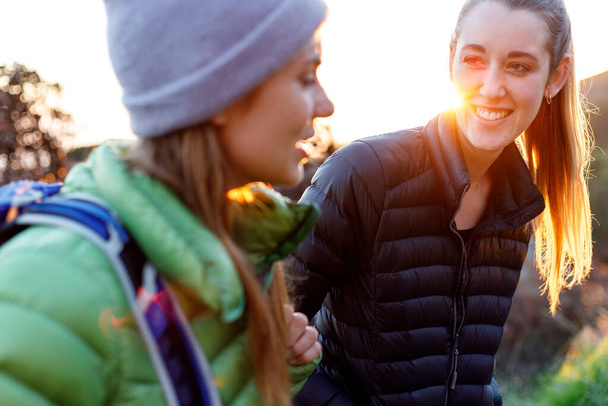 Smiling young woman in down jacket talking with female friend while hiking against clear sky during sunrise - Fotoğraf, Görsel