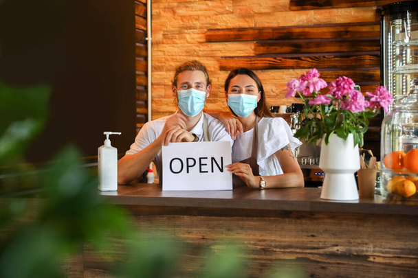 Власники бізнесу з медичними масками та відкриваючим знаком за столом у своєму кафе
 - Фото, зображення