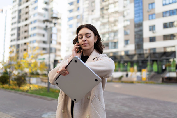 moderne succès jeune femme d'affaires parler sur un téléphone portable et en regardant sa montre avec un ordinateur portable dans ses mains. - Photo, image