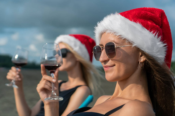 Портрет двох молодих жінок у купальниках, сонцезахисних окулярах і капелюхах Санта Клауса зі склянкою вина в руках сидять на складаних стільцях на піщаному узбережжі моря і насолоджуються моментом. - Фото, зображення
