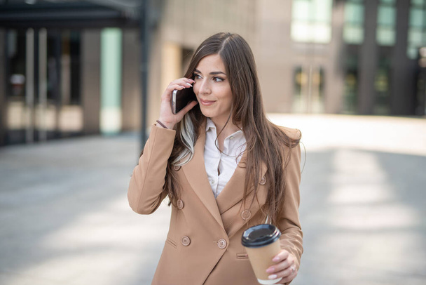 Young businesswoman talking on the phone in a city street while drinking coffee - Valokuva, kuva