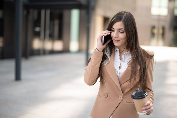 Jonge zakenvrouw praten aan de telefoon in een straat van de stad tijdens het drinken van koffie - Foto, afbeelding