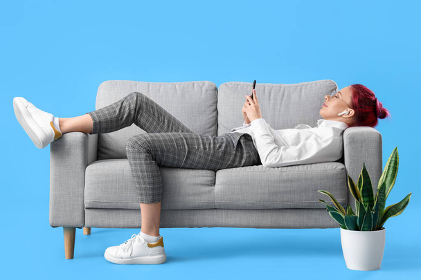 Pretty young woman with unusual hair and phone lying on sofa against blue background - Foto, Imagen