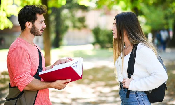 Two students, male and female, talking outdoor in a college park while holding book - Foto, afbeelding