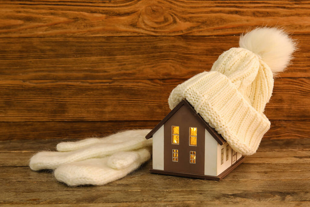 Modelo de casa con sombrero cálido y manoplas sobre fondo de madera oscura. Concepto de calefacción - Foto, Imagen