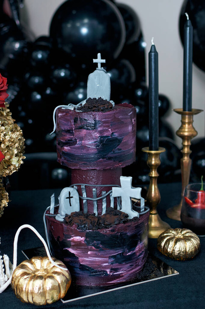  Hree többszintű halloween torta. A tortát sírkövekkel díszítették. Közel arany sütőtök és fekete gyertyák. Halloween desszert óra. - Fotó, kép