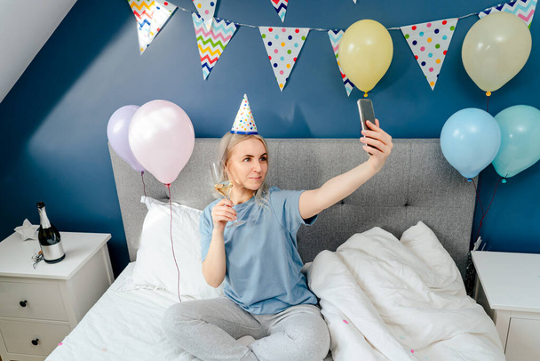 Glückliche emotionale Frau in Pyjama und Partymütze beim Selfie mit dem Handy am Geburtstagmorgen, während sie mit Champagner und Luftballons im Schlafzimmer auf dem Bett sitzt. Happy Birthday Konzept. - Foto, Bild