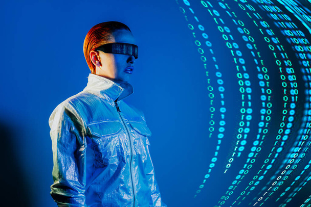 Woman, dressed in futuristic holographic clothes and eyeglasses with reflection of matrix code and matrix digital numbers artificial intelligence before her in blue light. Selective focus - Foto, afbeelding
