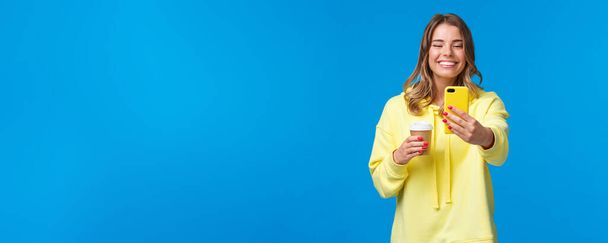 Joyful pretty feminine blond short-haired woman in yellow hoodie, taking selfie with take-away coffee from favorite local cafe, smiling smartphone camera standing with mobile phone. - Photo, Image