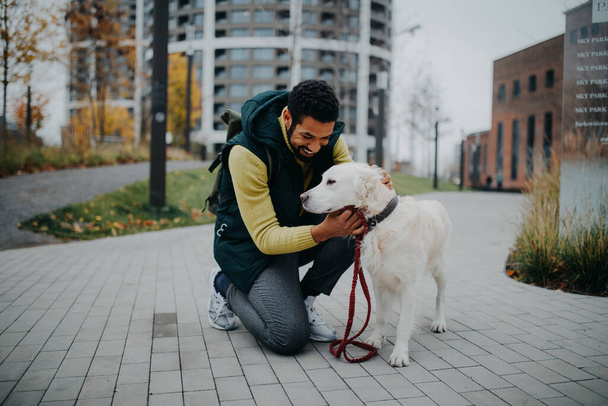 Happy young man stroking his dog outdoors in city park, during a cold autumn day. - Φωτογραφία, εικόνα