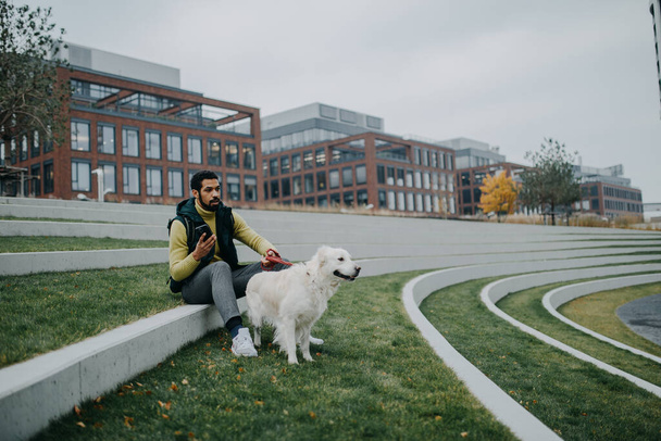 Happy young man resting and phoneing in urban city area during dog walk outdoors in cloudy autumn day. - Photo, Image