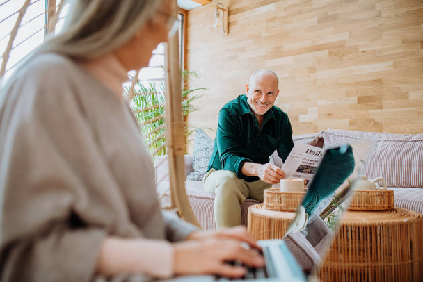 Senior woman sitting in indoor swing and working at laptop, her husband resting at a sofa. Concept of home office and active senior lifestyle. - Фото, изображение