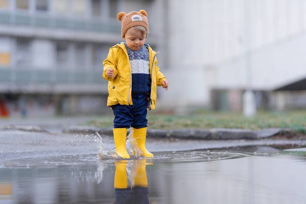 Happy little boy in yellow raincoat jumping in puddle after rain in cold autumn day. - Foto, Imagem