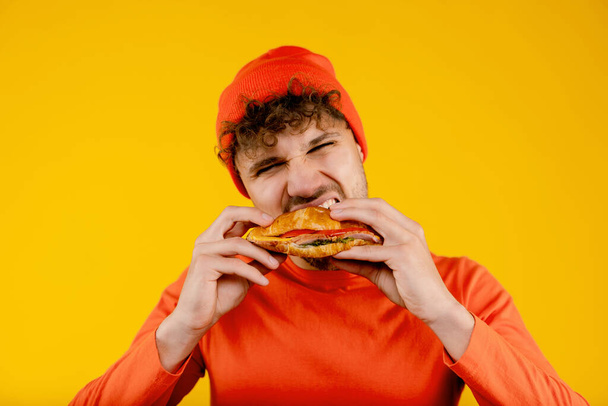 Joven hombre guapo hambriento muerde sándwich en ambas manos sobre un fondo amarillo aislado. Foto de alta calidad - Foto, Imagen