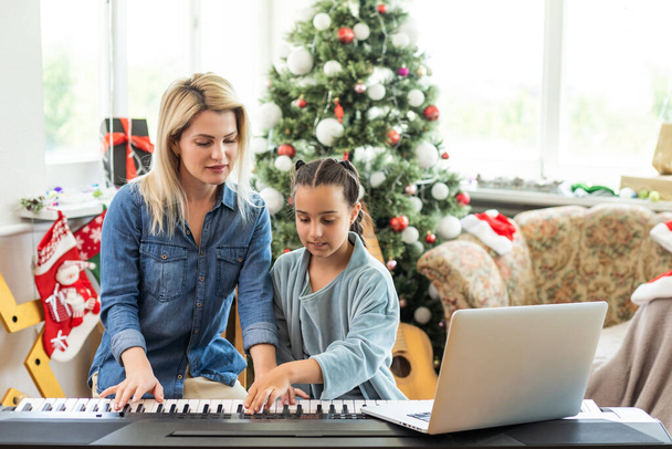 Merry Christmas and Happy Holidays. Cheerful mom and her cute daughter girl playing on a white piano against the background of a decorated Christmas tree. New Year. - Photo, Image