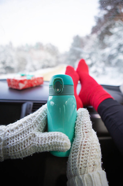 Una chica se sienta en calcetines rojos en un coche en invierno y se calienta con café de un termo, sobre el fondo de un bosque de invierno. Conceptos de viaje, viaje e invierno - Foto, imagen