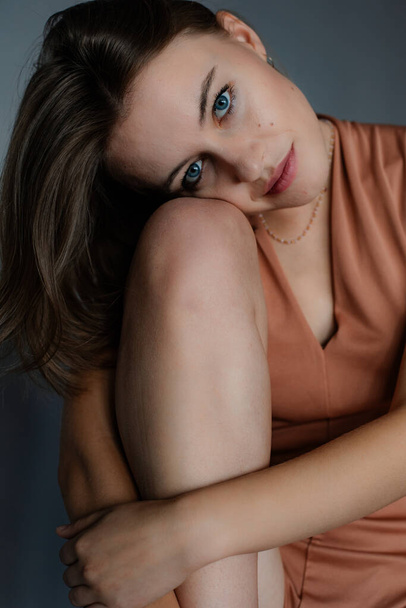 Portrait of young flawless blue-eyed woman with short dark hair wearing beige bodysuit sitting on floor, hugging knees on grey background, looking at camera. Beauty, bodycare, skincare, advertisement. - Fotoğraf, Görsel
