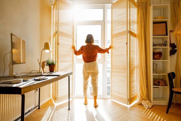 Woman opens window blinds letting the sun inside the room, spending good morning in sunny and cozy apartment in beige tones. Interior view - Foto, Imagem