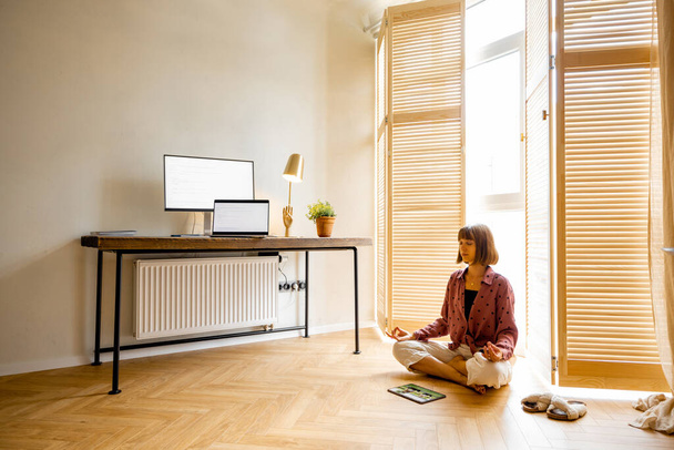 Mujer joven medita después del trabajo, sentado con la tableta en el suelo en la acogedora oficina en casa. Elegante interior con lugar de trabajo y ordenadores. Salud mental y relajación, trabajando desde el concepto del hogar - Foto, imagen