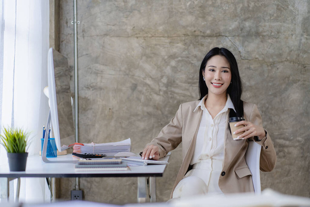 Donna d'affari asiatica che lavora su computer portatile con documento grafico finanziario in ufficio moderno vicino alla finestra con calcolatrice e rapporto d'affari sulla scrivania - Foto, immagini