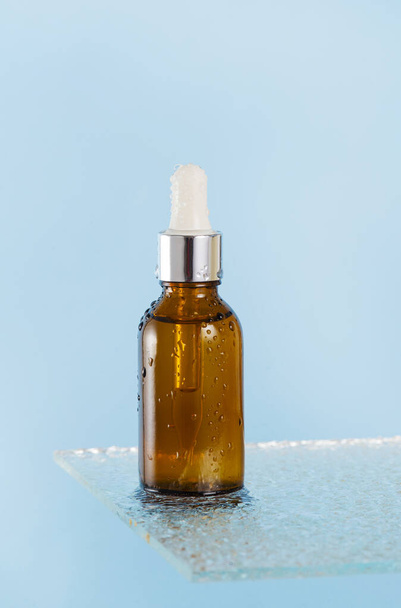 Een bruine druppelaar met een cosmetisch product, met een serum voor gezichtshuid op een glazen plank met waterspatten op blauwe achtergrond. - Foto, afbeelding