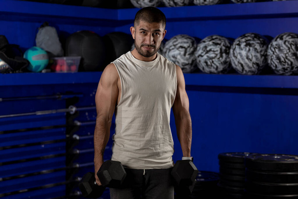 Латиноамериканец в спортивной форме в спортзале с гантелями в руках, смотрящий в камеру - Фото, изображение
