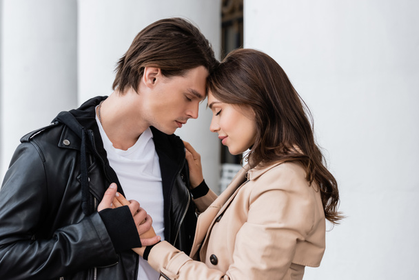 pleased young woman in beige trench coat hugging boyfriend in stylish jacket near white columns - Photo, Image