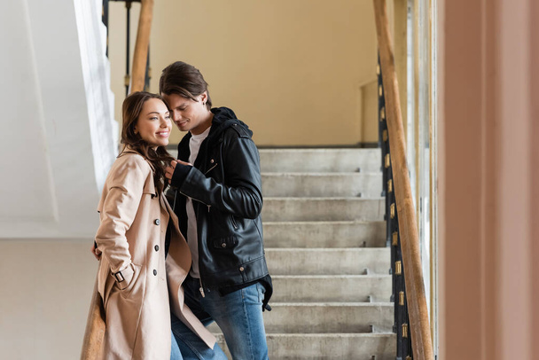 mladý muž ve stylové bundě flirtování s veselou přítelkyní v béžové trenč kabát stojící s rukou v kapse - Fotografie, Obrázek