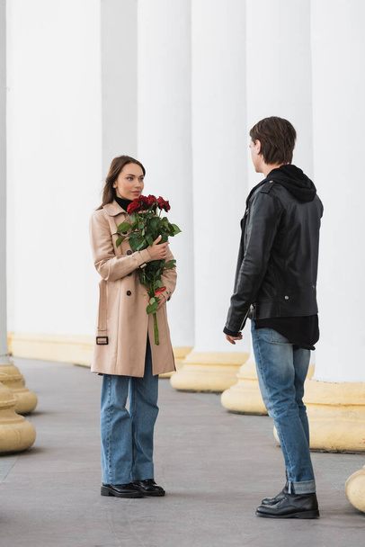 full length of pretty young woman holding red roses and looking at stylish boyfriend in jacket - Foto, immagini
