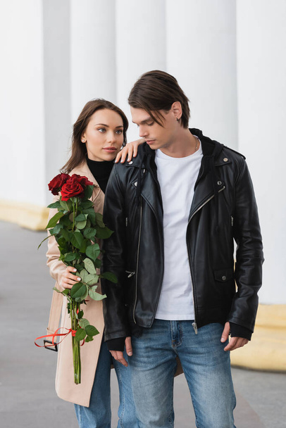 pretty young woman holding red roses and hugging shoulder of stylish boyfriend in jacket - Valokuva, kuva