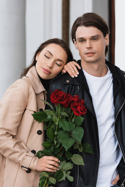 bella donna in trench in possesso di rose rosse e appoggiata sulla spalla del fidanzato in giacca elegante - Foto, immagini