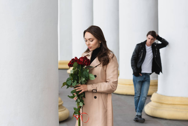pretty young woman in beige trench coat looking at red roses near boyfriend on blurred background  - Photo, Image
