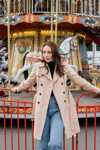pretty young woman in beige trench coat and jeans standing near carousel in amusement park - Foto, Imagem