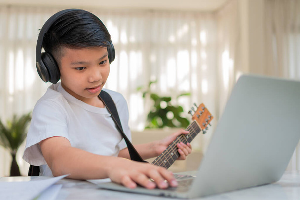 Asijský chlapec hraje na kytaru a sledovat on-line kurz na notebooku při tréninku pro učení hudby nebo hudebního nástroje on-line doma. Chlapec studenti studovat online s video call učitelé hrát na kytaru. - Fotografie, Obrázek