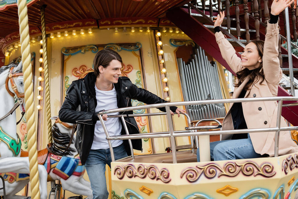 cheerful man in trendy outfit looking at happy girlfriend with raised hands on carousel in amusement park - 写真・画像