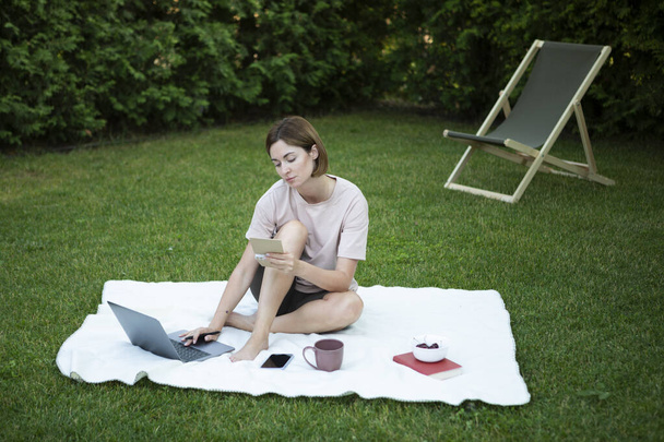 Привлекательная молодая женщина, сидящая на лужайке, работающая над ноутбуком и держащая в руке блокнот для записи творческих идей. Работа и отдых одновременно концепция - Фото, изображение
