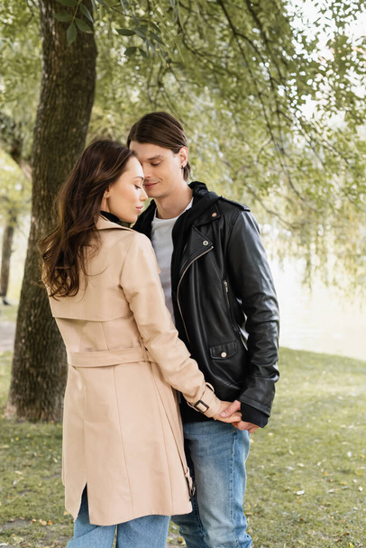pretty young woman in trench coat and cheerful man in jacket holding hands in park - Foto, Imagen