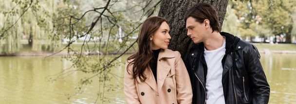 stylish man and pretty woman in trench coat looking at each other near river in park, banner - Photo, Image