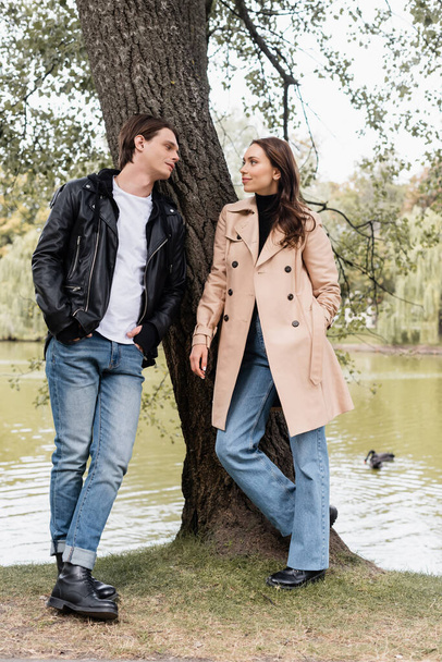 volledige lengte van stijlvolle man en mooie vrouw in trench jas kijken naar elkaar in de buurt van rivier in park - Foto, afbeelding