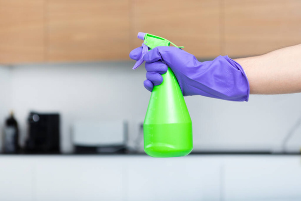 Primer plano de la mano en guante violeta con spray de botella verde para limpiar el fondo de la cocina. Concepto de servicio doméstico - Foto, Imagen