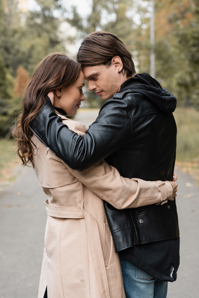 Seitenansicht des stilvollen jungen Mannes umarmt hübsche Freundin im Trenchcoat im Freien - Foto, Bild