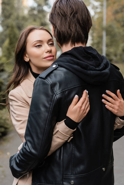 junge und hübsche Frau im beigen Mantel umarmt Freund in schwarzer Jacke  - Foto, Bild