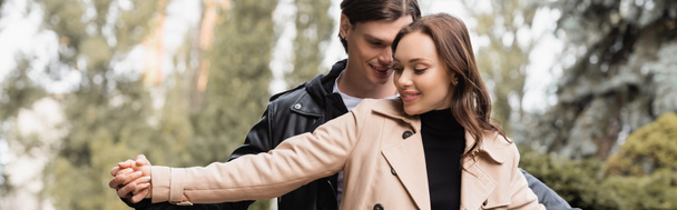vrolijke man hand in hand met tevreden vriendin in beige trench jas in park, banner - Foto, afbeelding
