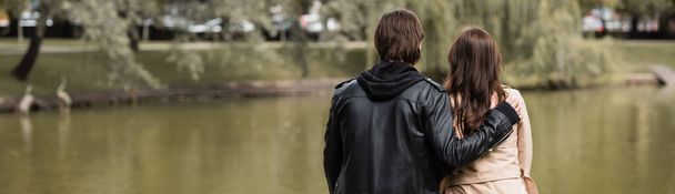 back view of young man in black jacket hugging girlfriend near lake in park, banner - Photo, Image