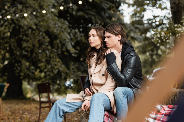 stylish man in jacket hugging young girlfriend in beige coat sitting in park - Zdjęcie, obraz