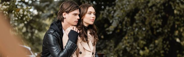 stylish man in jacket hugging young girlfriend in beige coat sitting in park, banner - Photo, image