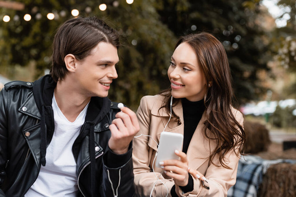 cheerful young woman holding smartphone while listening music and sharing wired earphones with happy boyfriend  - Fotografie, Obrázek