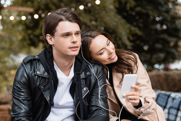 cheerful woman holding smartphone while sharing wired earphones with boyfriend and listening music together - Photo, Image