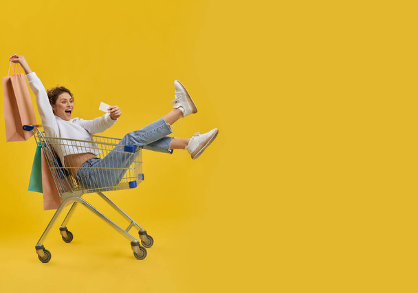 Side view of pretty girl driving in shopping cart. Pretty young woman sitting, shouting, smiling, taking selfie, holding packages, raising hand. Concept of carefree youth life. - Foto, immagini