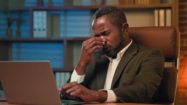 Hombre de negocios afroamericano adulto que trabaja en el ordenador portátil en la oficina hombre cansado poco saludable que sufre de dolor de cabeza siente dolor presión fatiga por exceso de trabajo sufren migraña crónica que experimenta problemas de salud - Imágenes, Vídeo